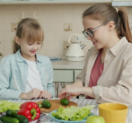 Young woman is cooking salad and feeding cute little daughter with cucumber slices in kitchen, family is talking and having good time together.