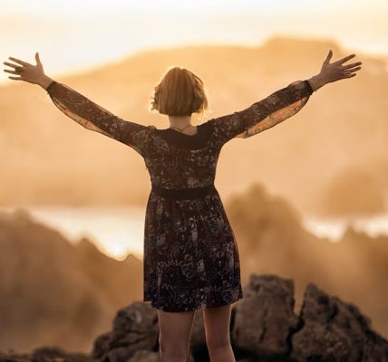 A woman stands with open arms on a mountain at sunset, embracing nature's beauty.
